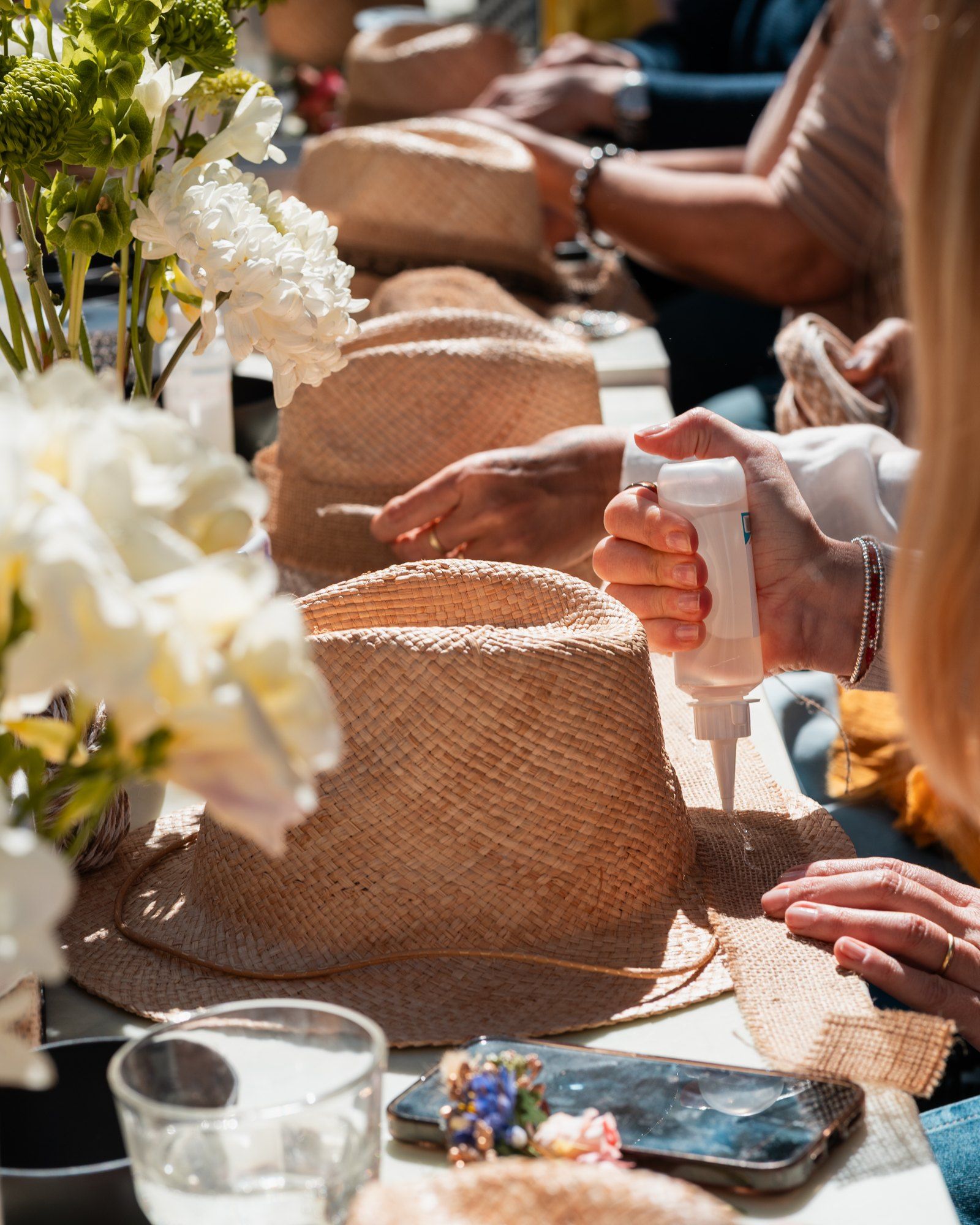 Manos que aplican pegamento sobre un sombrero de paja, entre flores blancas.