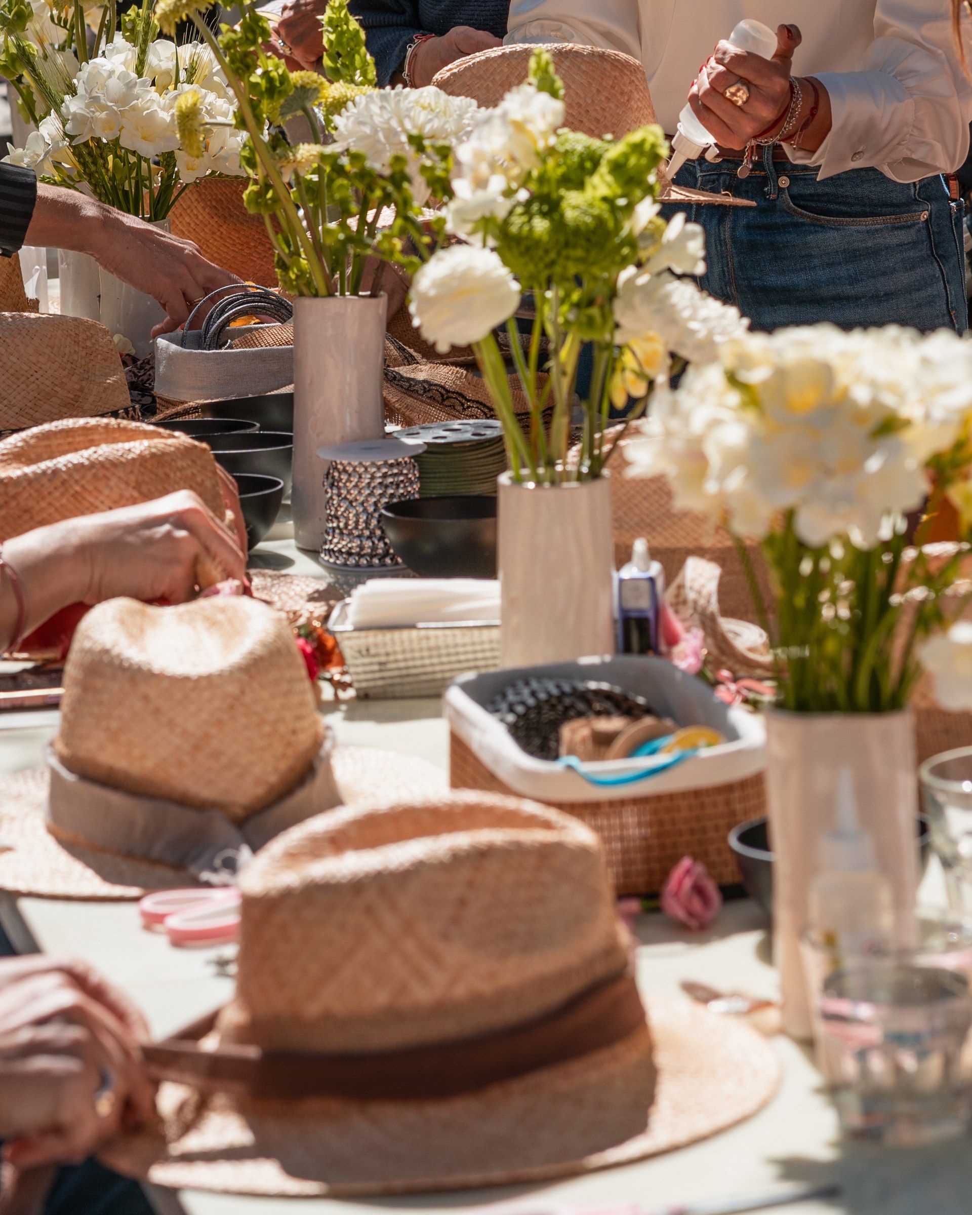 Mesa de trabajo con sombreros de paja a medio intervenir, flores blancas y avíos.