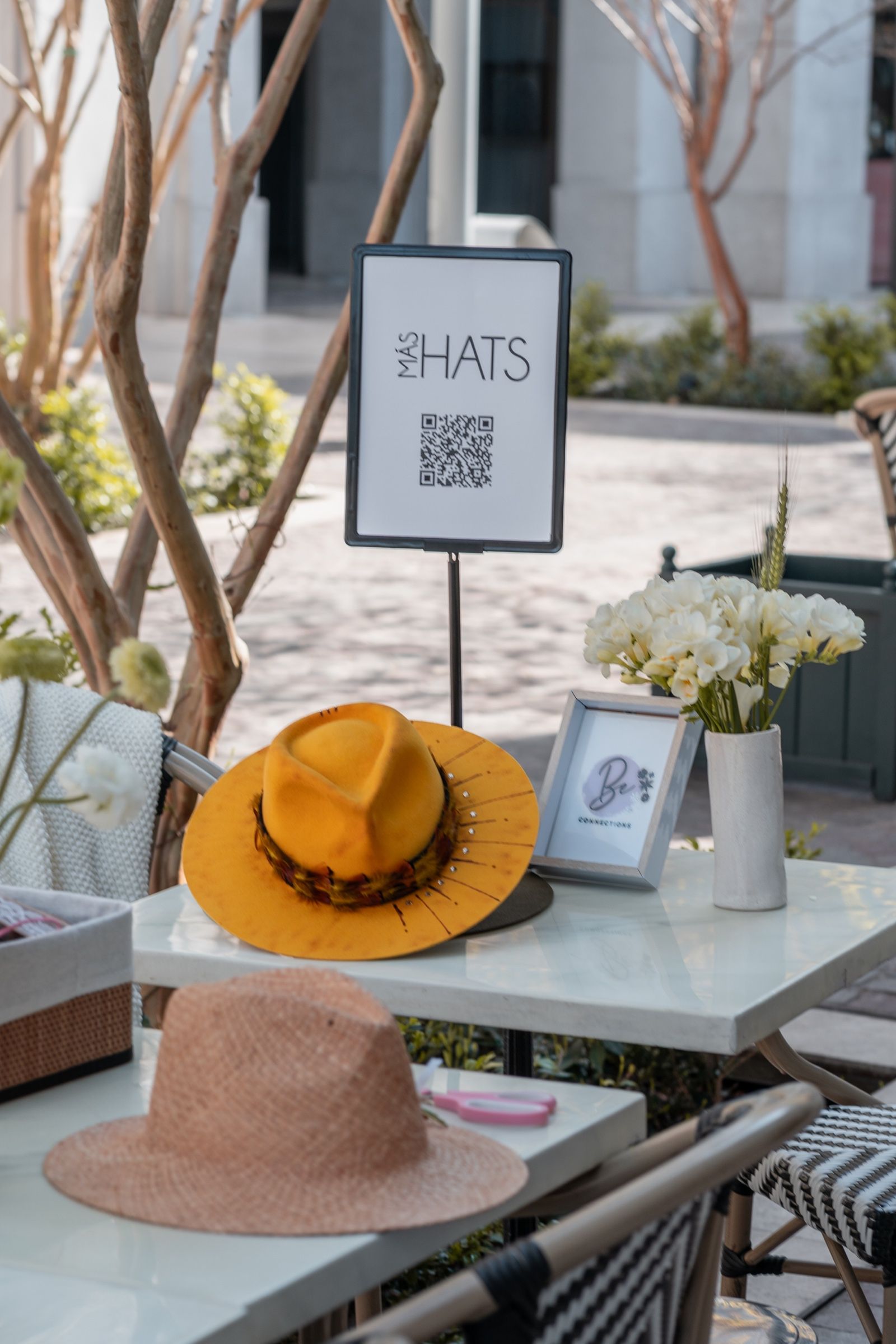 Mesa preparada con un sombrero amarillo, un sombrero de paja y el cartel de MÁS HATS.