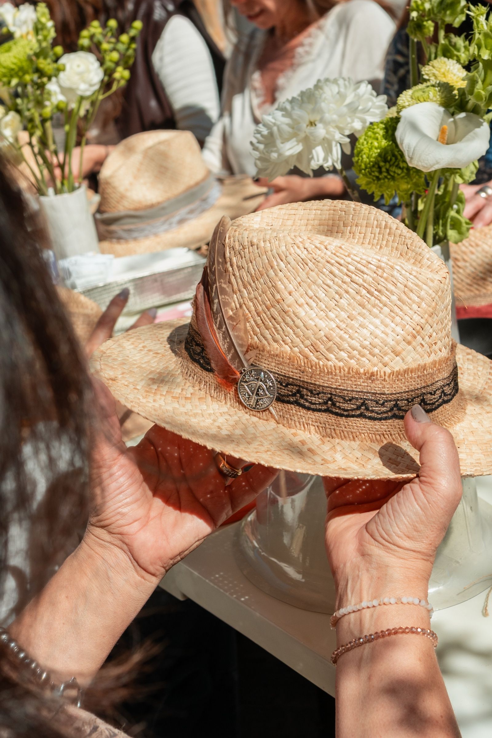 Manos que sostienen un sombrero de paja terminado, con cinta bordada y una medalla.